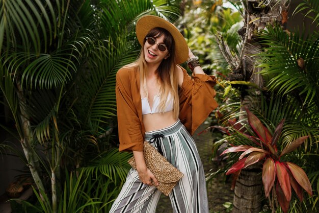 Imagen de moda de mujer elegante sexy con sombrero de paja posando en hojas de palmeras tropicales