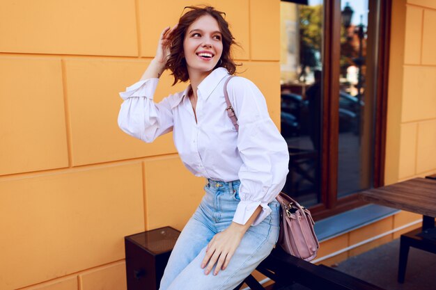 Imagen de moda de niña bonita morena con peinado corto en elegante blusa blanca casual y jeans. Botas de cuero negro con tacones. Chica sentada cerca de café moderno con paredes amarillas.