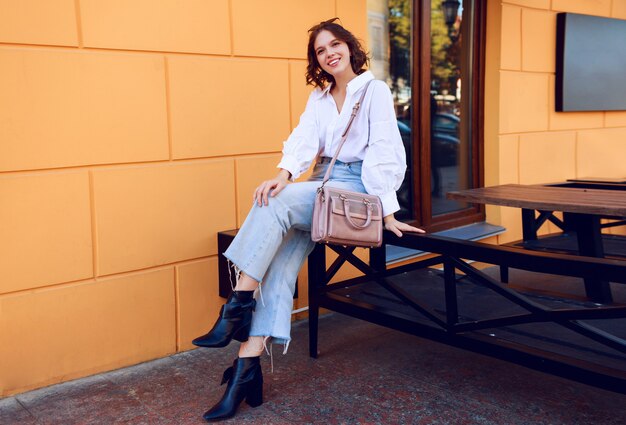 Imagen de moda de niña bonita morena con peinado corto en elegante blusa blanca casual y jeans. Botas de cuero negro con tacones. Chica sentada cerca de café moderno con paredes amarillas.