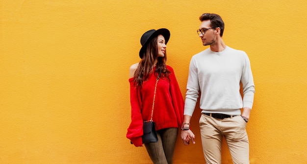 Foto gratuita imagen de moda otoño de elegante elegante pareja de enamorados cogidos de la mano y mirando el uno al otro con placer. mujer de pelo largo en suéter de punto rojo con su novio posando