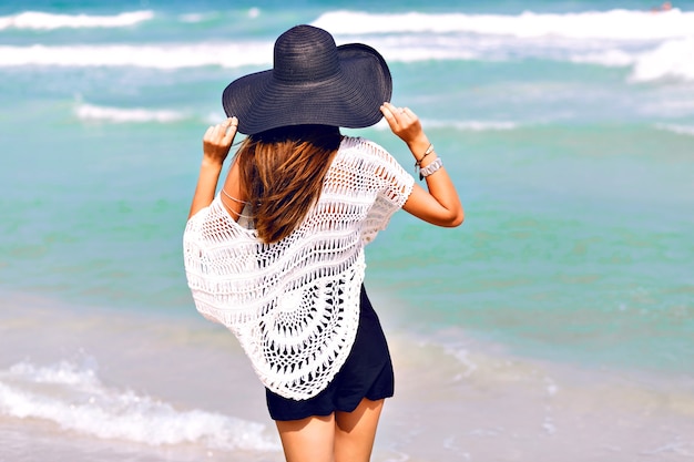 Imagen de moda de verano de mujer posando de nuevo, cerca del agua de mar azul, agradable día soleado de verano, relajarse y disfrutar de la libertad, alegría, felicidad, colores brillantes