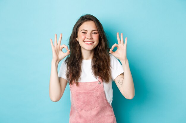 La imagen de la modelo femenina morena sonriente dice que sí, muestra signos de aprobación, está de acuerdo o elogia la buena elección, recomienda el producto, de pie contra el fondo azul.