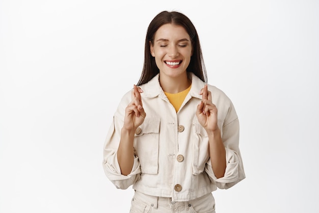 Imagen de una modelo femenina sonriente y esperanzada que cierra los ojos con los dedos cruzados y pide un deseo anticipando buenas noticias saborear un resultado positivo fondo blanco Copiar espacio