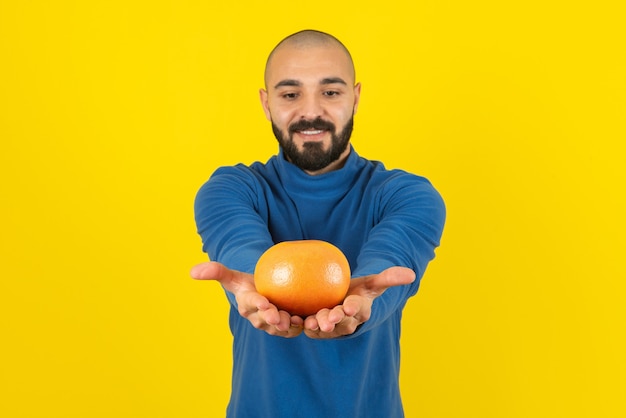 Imagen de un modelo de hombre mostrando una fruta naranja contra una pared amarilla.