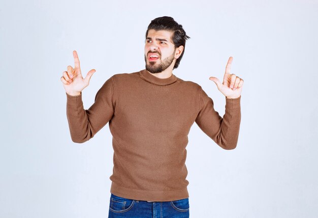Imagen de modelo masculino joven en suéter marrón de pie sobre una pared blanca.