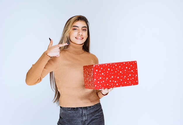 Imagen de un modelo de mujer atractiva sosteniendo un regalo rojo y apuntando hacia otro lado.