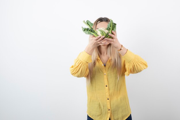 Imagen de un modelo de mujer joven y bonita de pie y cubriendo su rostro con coliflor.