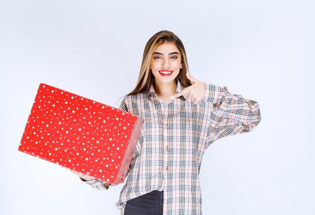 Imagen de un modelo de mujer joven de pie y apuntando a una caja de regalo roja.