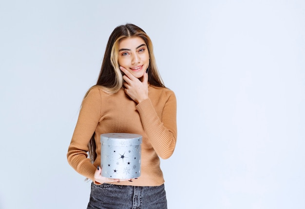 Imagen de una modelo de mujer joven en suéter marrón de pie y posando con una caja de regalo.