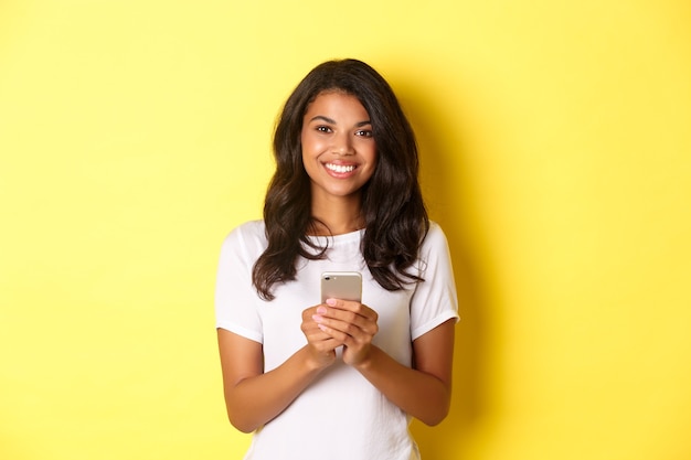 Imagen de la moderna niña afroamericana sonriendo con teléfono móvil de pie sobre fondo amarillo