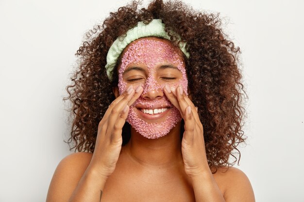Imagen de una mujer afroamericana bonita feliz que se pela la cara con un exfoliante de sal marina rosa, toca las mejillas, se para los hombros desnudos contra la pared blanca. Concepto de belleza y cuidado personal.