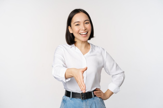 Imagen de una mujer asiática confiada sonriendo extendiendo la mano para un gesto de saludo de apretón de manos diciendo hola de pie sobre fondo blanco