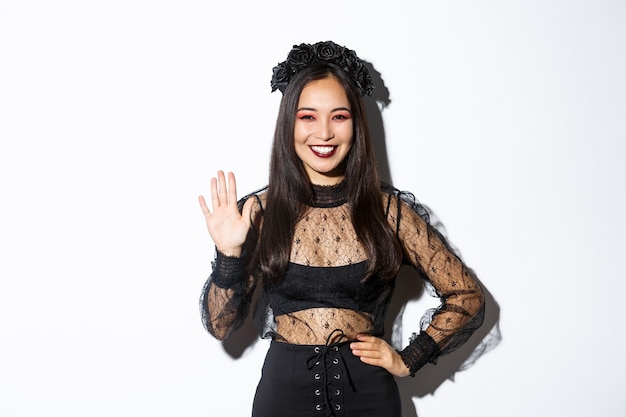 Imagen de mujer asiática elegante y amistosa en vestido de encaje gótico agitando la mano para saludar, saludar a alguien, dar la bienvenida a la gente en la fiesta de Halloween, de pie sobre fondo blanco.