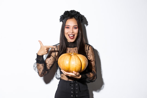 Imagen de mujer asiática emocionada con maquillaje gótico, vestido de bruja negro y sosteniendo calabaza, de pie asombrado sobre fondo blanco.