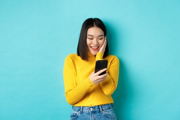 Imagen de la mujer asiática feliz leyendo un mensaje en la pantalla del teléfono móvil y sonriendo, charla en la aplicación del teléfono inteligente, de pie sobre fondo azul