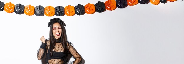 Imagen de mujer asiática feliz en traje de bruja divirtiéndose en la fiesta de halloween diciendo sí standi alegre