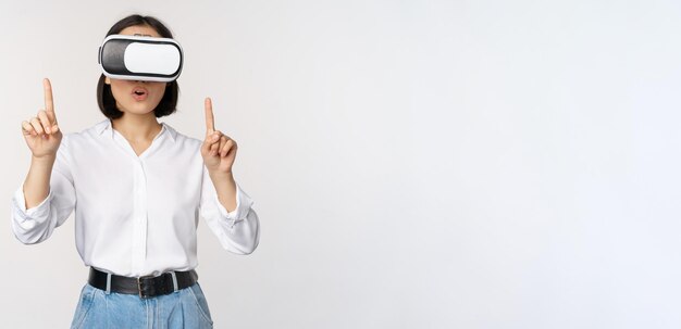 Imagen de una mujer asiática con gafas vr apuntando con el dedo hacia arriba mirando a una chica asombrada y sorprendida usando virtu