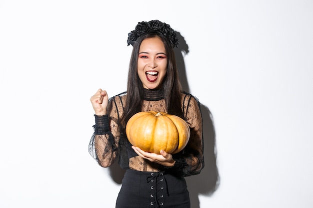 Imagen de mujer asiática hermosa emocionada celebrando halloween, vistiendo traje de bruja y sosteniendo calabaza, gritando de alegría.