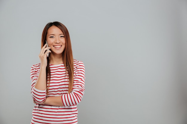 Imagen de la mujer asiática joven feliz que habla por la situación del teléfono aislada sobre la pared gris. Mirando la cámara.