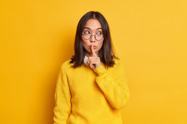 Imagen de mujer asiática misteriosa que hace gesto de silencio comparte miradas secretas a un lado tiene cabello oscuro y apariencia específica vestida con cálido jersey.