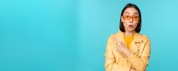 La imagen de una mujer asiática parece intrigada hace una pregunta sobre el artículo o la tienda señala con el dedo a la izquierda con una expresión de cara sorprendida sobre fondo azul