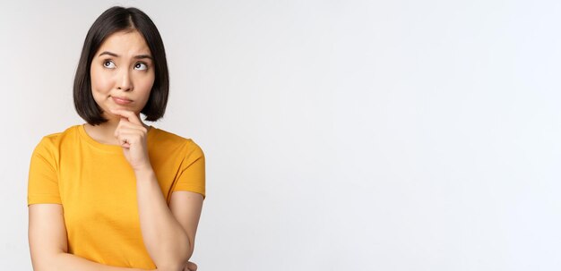 Imagen de una mujer asiática pensante mirando a un lado y reflexionando sobre la toma de decisiones de pie en una camiseta amarilla sobre fondo blanco.