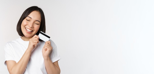 Imagen de una mujer asiática sonriente abrazando con tarjeta de crédito comprando sin contacto de pie en camiseta blanca sobre wh