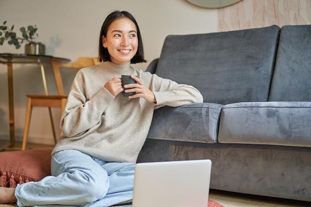 Imagen de una mujer asiática sonriente bebiendo té caliente sosteniendo una taza y sentada cerca de una laptop en el piso descansando un