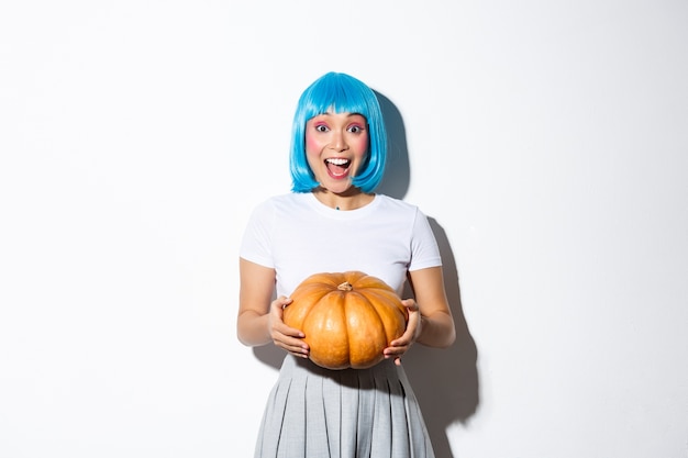 Imagen de mujer asiática sonriente emocionada celebrando halloween, sosteniendo una gran calabaza, con peluca azul para la fiesta, de pie.