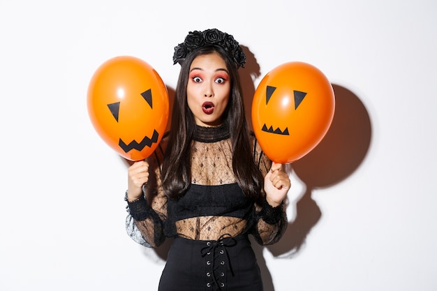 Imagen de mujer asiática sorprendida en traje de bruja celebrando halloween, sosteniendo globos con caras aterradoras, de pie sobre fondo blanco.