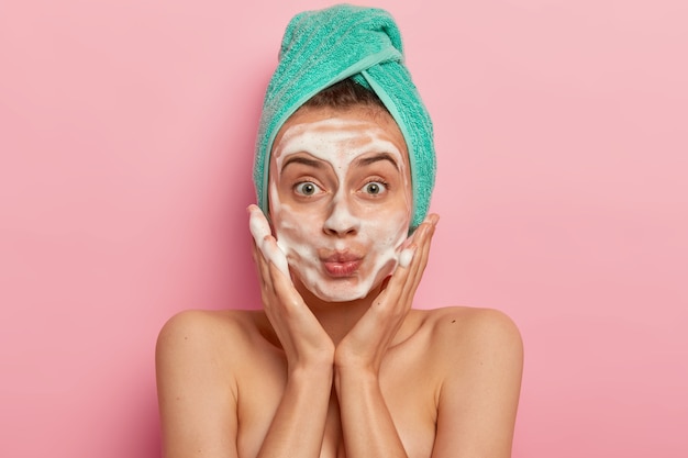 Foto gratuita imagen de mujer atractiva que se lava la cara con espuma, masajea las mejillas, se mira sorprendentemente a sí misma, usa una toalla envuelta en la cabeza, elimina la suciedad, siente frescura después de tomar una ducha, modelos en interiores