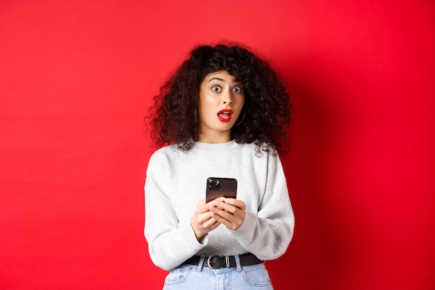 Imagen de mujer bastante rizada con cara de sorpresa, leyendo el mensaje en el teléfono inteligente sorprendido, de pie contra el fondo rojo.