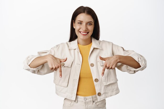 Imagen de una mujer cándida sonriente, expresión facial agradable y amistosa, señalando con el dedo hacia abajo, mostrando un anuncio de venta debajo, de pie sobre fondo blanco