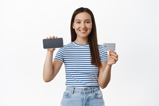 Imagen de una mujer caucásica atractiva que muestra una pantalla de teléfono inteligente horizontal, teléfono móvil volteado y tarjeta de crédito, concepto de publicidad, fondo blanco