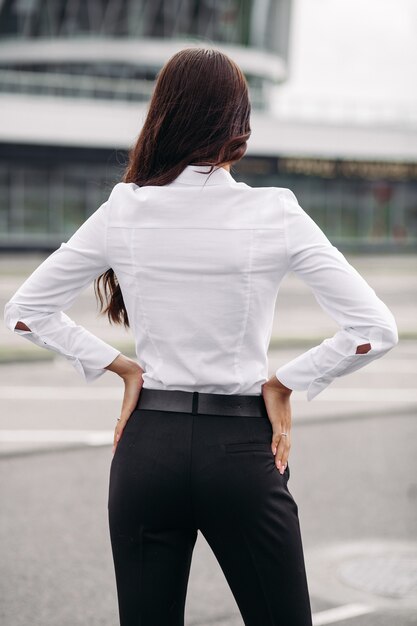 Imagen de una mujer caucásica hermosa con el pelo largo y ondulado oscuro con una camisa blanca, pantalón negro y tacones mira edificio alto