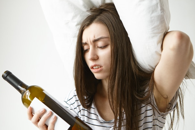 Imagen de una mujer caucásica joven estresada con el ceño fruncido en pijama de rayas sosteniendo la almohada en la cabeza y mirando la botella vacía, sintiéndose desesperada cuando el vino se acabó, sufriendo de resaca después de la fiesta