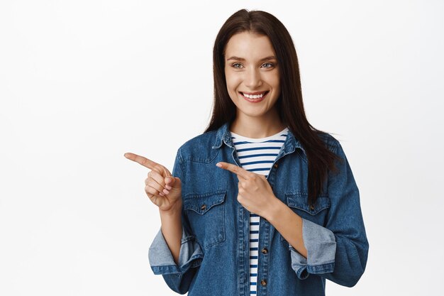 Imagen de una mujer caucásica señalando, mirando a la izquierda con una sonrisa satisfecha, una clienta señala el producto que quiere, recogiendo algo en la tienda, de pie contra un fondo blanco.