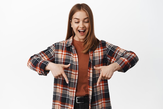 Imagen de una mujer caucásica sonriente mirando, apuntando hacia abajo con una expresión de cara feliz asombrada, mostrando un nuevo anuncio genial, producto de logotipo, fondo blanco.