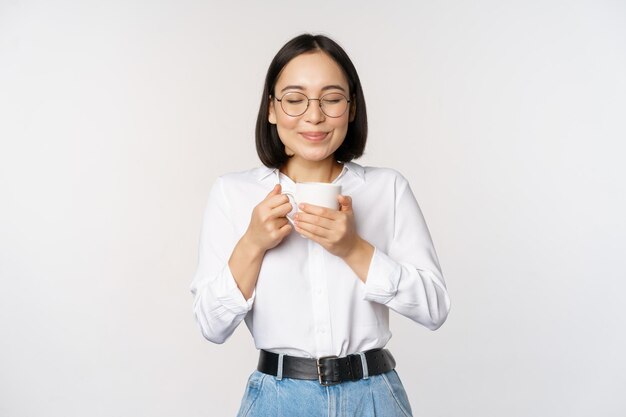 Imagen de una mujer coreana sonriente oliendo café en una taza disfrutando de una deliciosa bebida de pie sobre fondo blanco