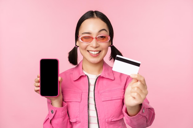 Imagen de una mujer coreana sonriente que muestra la tarjeta de crédito y la interfaz de la aplicación del teléfono inteligente de la pantalla del teléfono móvil pagando compras en línea sin contacto de pie sobre fondo rosa