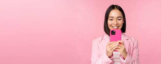 Imagen de una mujer corporativa asiática sonriente con traje mirando mirando en la aplicación de teléfono inteligente usando la aplicación de teléfono móvil de pie sobre fondo rosa