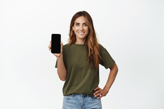 Imagen de una mujer elegante y sonriente que muestra la pantalla, la pantalla móvil en blanco, la interfaz del teléfono, la aplicación recomendada, el anuncio celular 4g, de pie sobre fondo blanco.