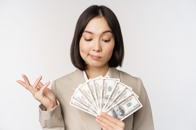 Imagen de mujer empresaria asiática empresaria feliz mostrando dinero en efectivo dólares y pensando de pie en traje sobre fondo blanco.