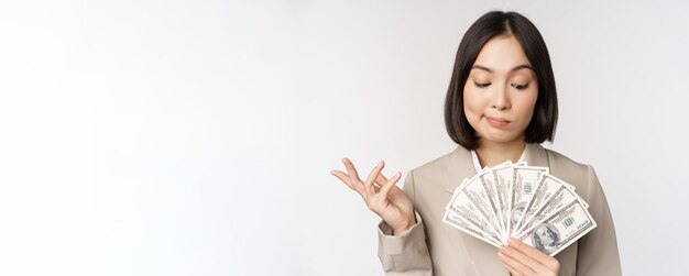 Imagen de mujer empresaria asiática empresaria feliz mostrando dinero en efectivo dólares y pensando de pie en traje sobre fondo blanco.