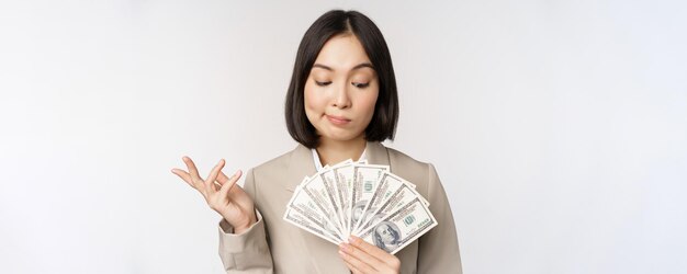 Imagen de mujer empresaria asiática empresaria feliz mostrando dinero en efectivo dólares y pensando de pie en traje sobre fondo blanco.