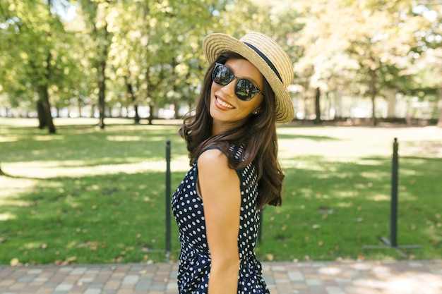 Imagen de mujer de estilo encantador está caminando en el parque de verano con sombrero de verano y gafas de sol negras y un lindo vestido.