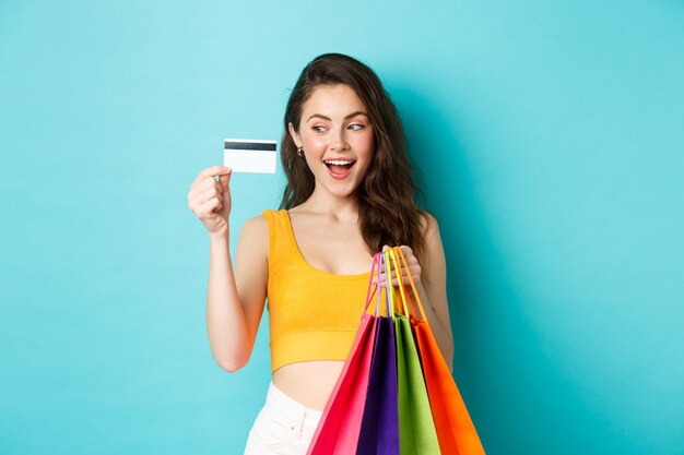 Imagen de mujer feliz adicta a las compras mostrando su tarjeta de crédito de plástico, sosteniendo bolsas de la compra, vistiendo ropa de verano, de pie contra el fondo azul.