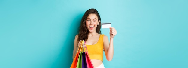 Foto gratuita imagen de una mujer feliz adicta a las compras que muestra su tarjeta de crédito de plástico sosteniendo bolsas de compras con ropa de verano de pie contra un fondo azul