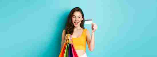 Foto gratuita imagen de una mujer feliz adicta a las compras que muestra su tarjeta de crédito de plástico sosteniendo bolsas de compras con ropa de verano de pie contra un fondo azul