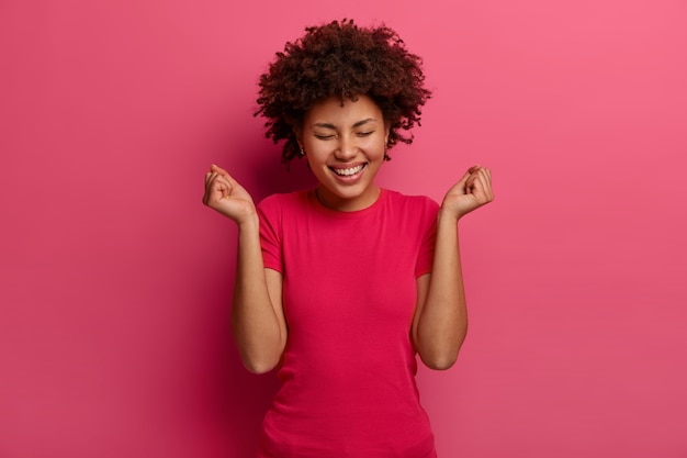 La imagen de una mujer feliz hace un gesto de bomba de puño, celebra una gran noticia, se ríe positivamente, viste ropa informal, se ríe positivamente, posa contra la pared rosa brillante. Emociones y concepto de éxito